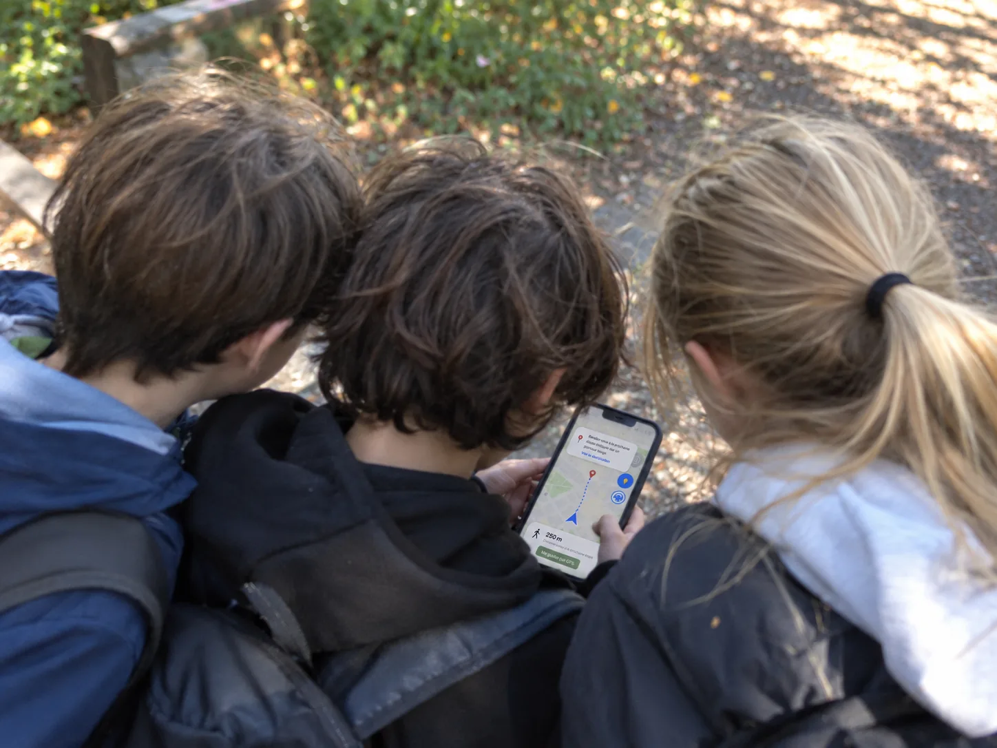 Trois élèves penchés sur un téléphone qui affiche l'itinéraire vers la prochaine étape, pendant une sortie en extérieur
