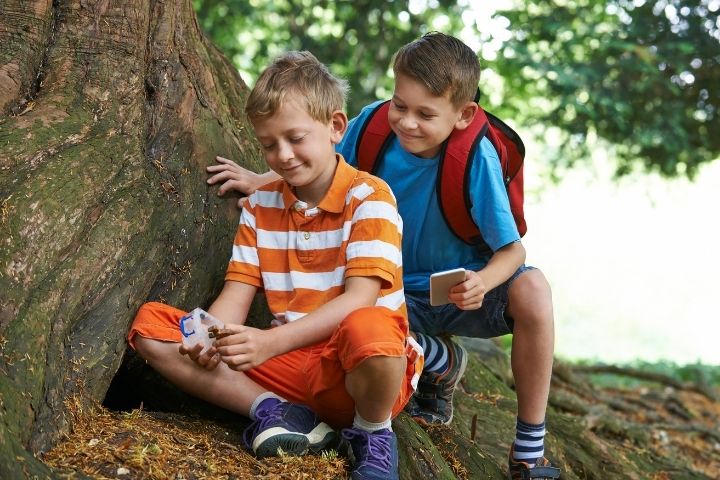 Deux enfants trouvent un indice au pied d'un arbre pendant un jeu de piste, téléphone en main